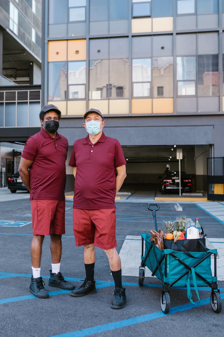 Polo Shirts Two delivery men in masks stand with a cart filled with groceries outdoors in a city.