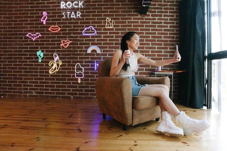Breckie Hill Nude Young woman in stylish casual wear taking a selfie in a chic room with neon decor and brick walls.
