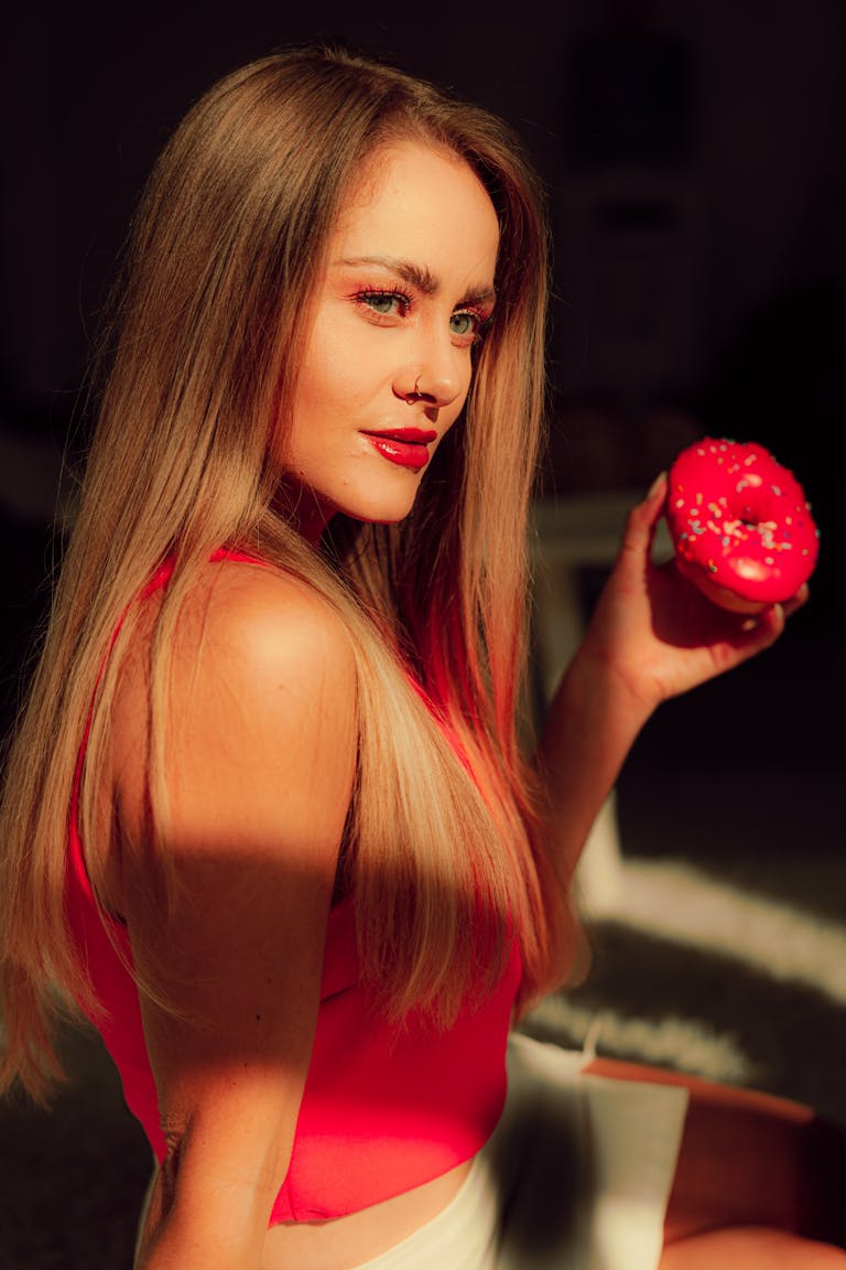 Candizi Beautiful woman in vibrant red top holds a pink donut, basking in warm sunlight.