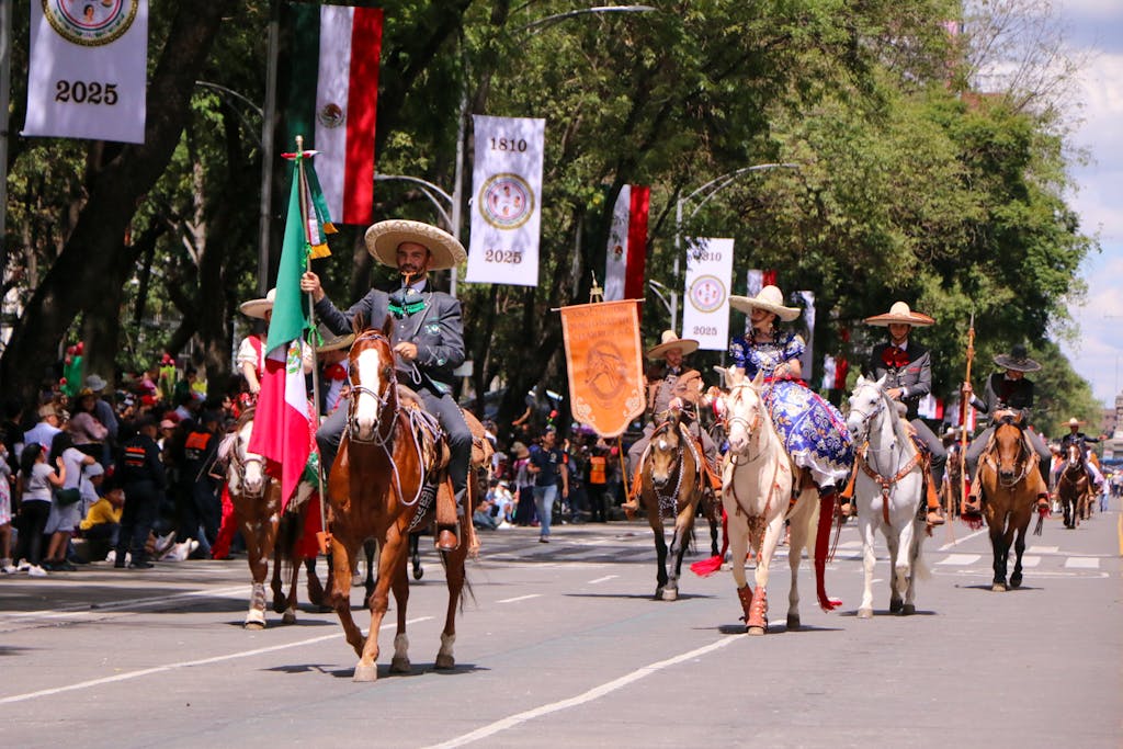 5 Movierulz 2025 Charros riding horses in a Mexican Independence Day parade with vibrant banners and flags.