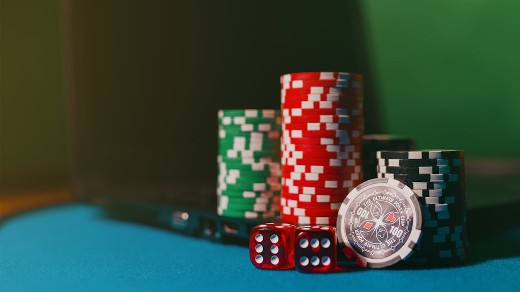 7700bet11 Close-up of casino chips and dice on a felt table, next to a laptop for online gambling.