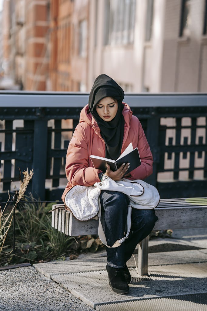 Full body of young Middle East female in pink jacket and black Islamic headscarf sitting on bench and reading novel on street