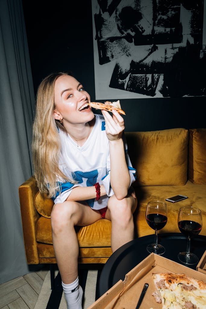 A happy woman in a casual setting enjoying a slice of pizza with red wine indoors.