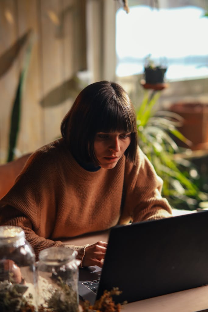 A woman in a cozy sweater concentrates on her laptop in a sunlit room, surrounded by plants.