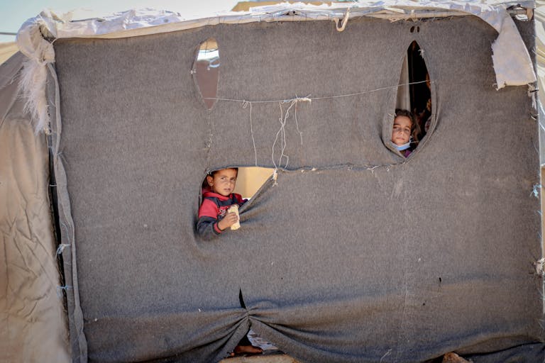 World War 2 Log: Children peeking through a makeshift tent in Idlib, highlighting resilience and hope.