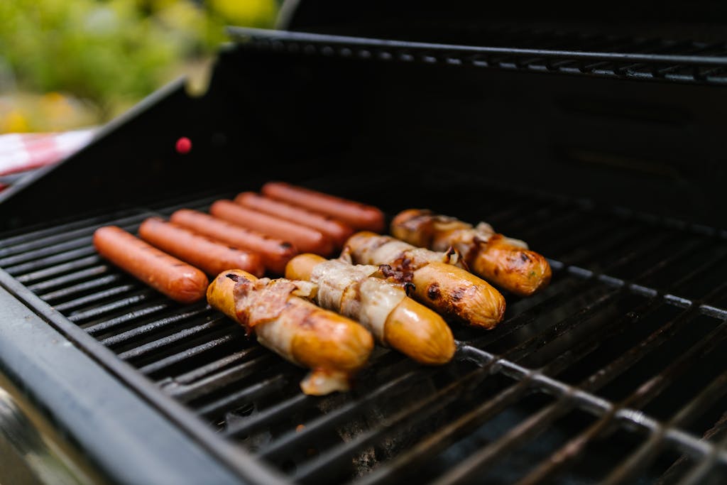 Weber Grillvorführung: Close-up of hotdogs and bacon-wrapped sausages on a grill outdoors.