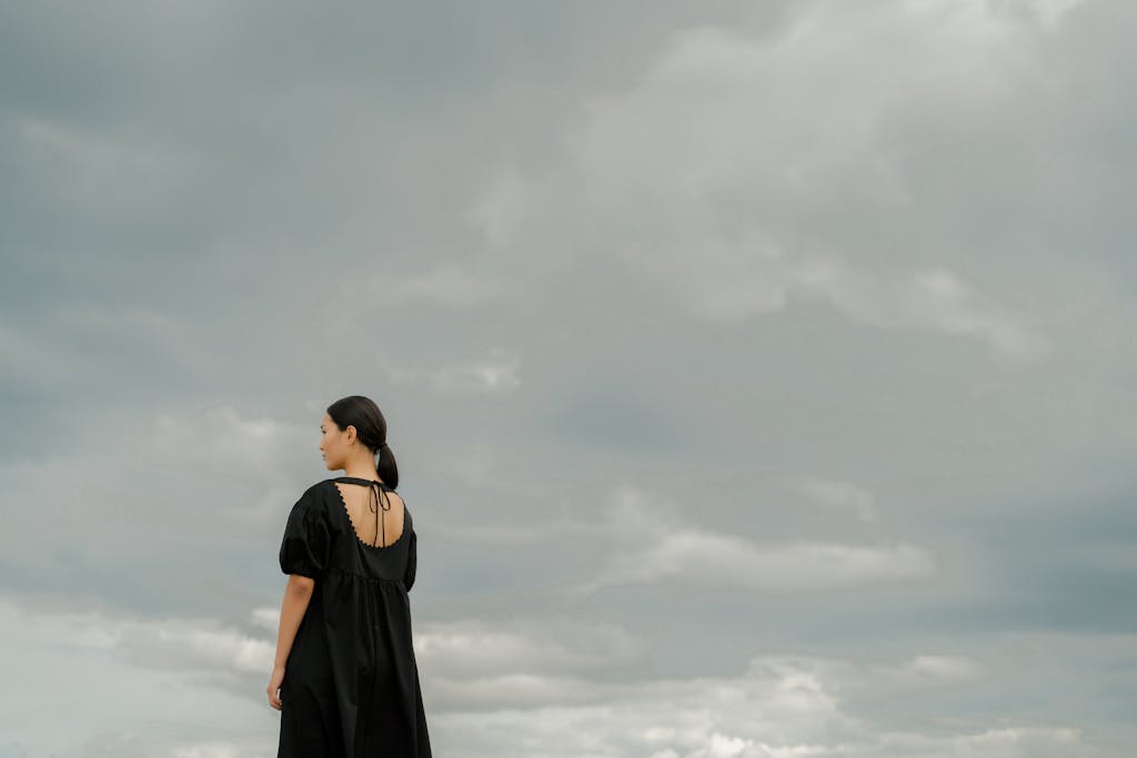 Carmela ClouthA woman in a black dress stands alone outdoors, gazing at a dramatic cloudy sky.